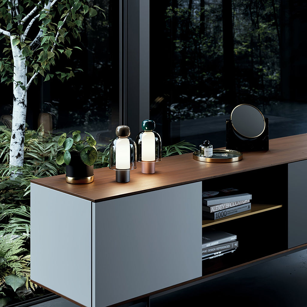 Two Clérachic table lamps lit on a pale cabinet with a wood top, with birch trunks and ferns visible through the window behind.