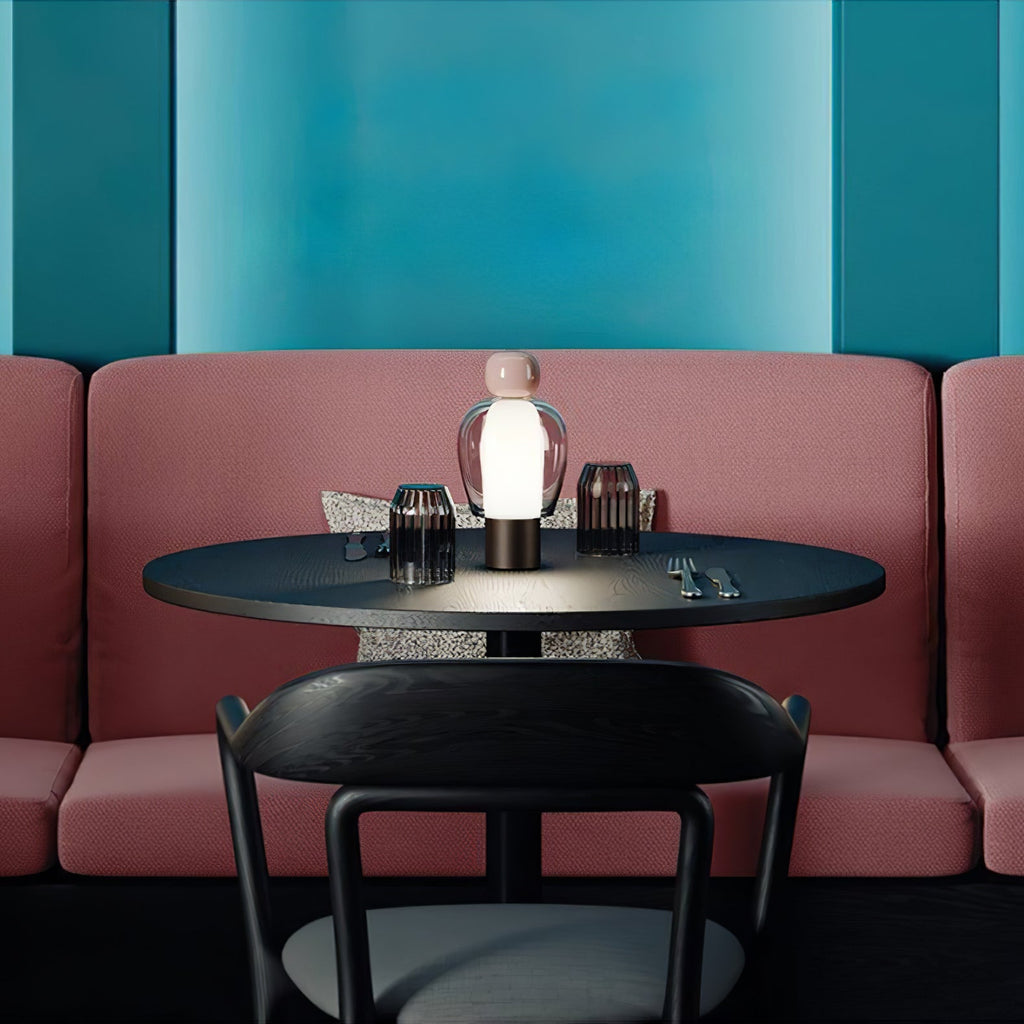 A single Clérachic table lamp glowing on a small black café table, with a pink banquette and teal wall behind.