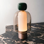 A Clérachic table lamp with a dark green cap and smoked glass dome glowing on black marble veined with gold, beside a pale wall.