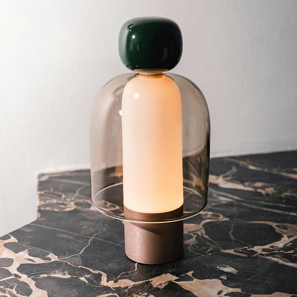 A Clérachic table lamp with a dark green cap and smoked glass dome glowing on black marble veined with gold, beside a pale wall.