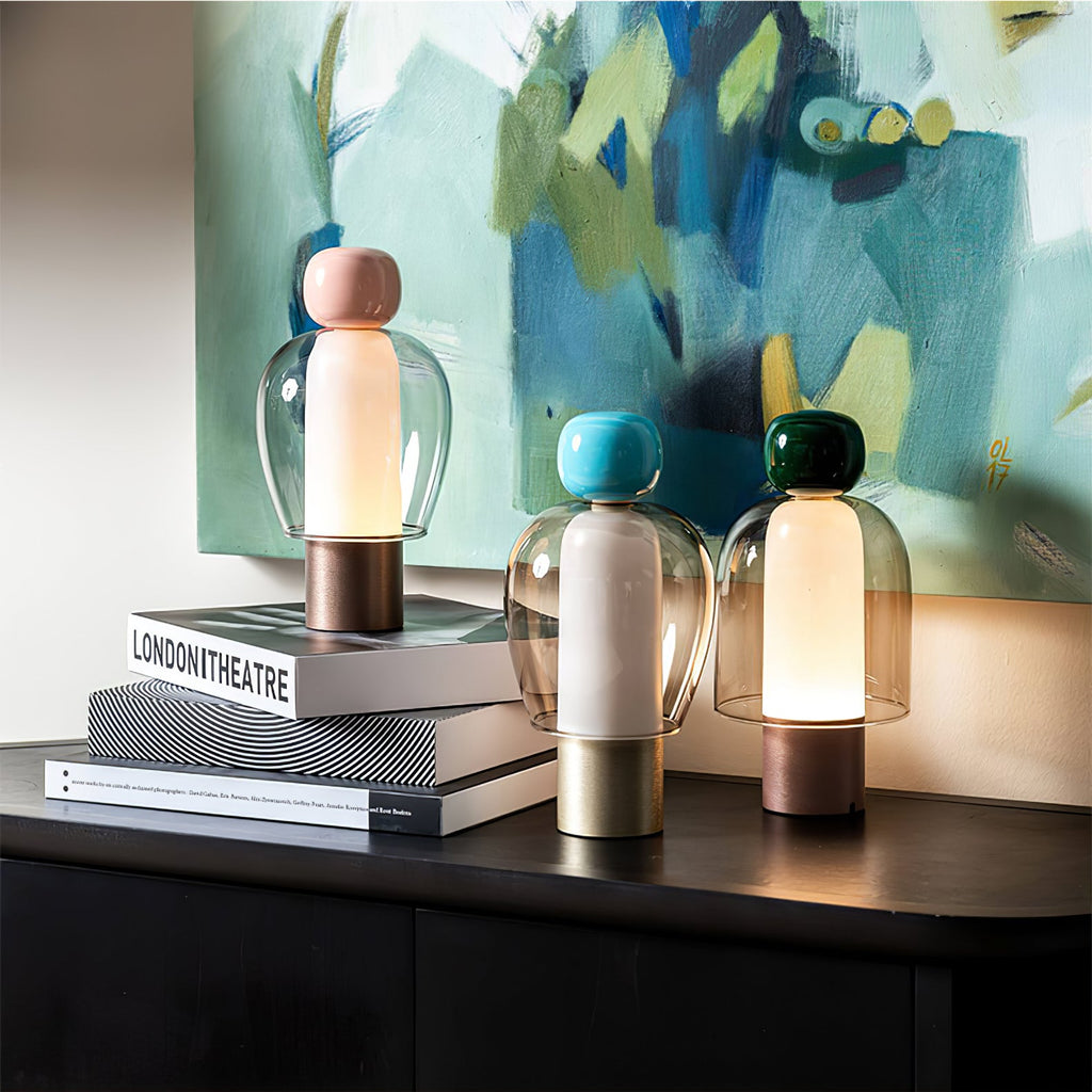 Three Clérachic table lamps in a row on a black cabinet, the nearest lit, with a stack of books and a blue-green painting behind.