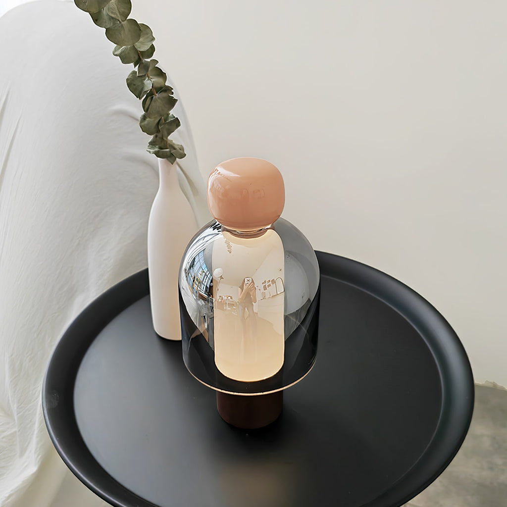 A Clérachic table lamp with a pink cap and smoked glass dome on a round black tray, beside a white vase holding a eucalyptus stem.
