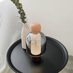 A Clérachic table lamp with a pink cap and smoked glass dome on a round black tray, beside a white vase holding a eucalyptus stem.