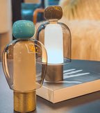 Close view of a turquoise-capped Clérachic table lamp in front of a lit brown-capped one, on a book with a gold-toned edge.