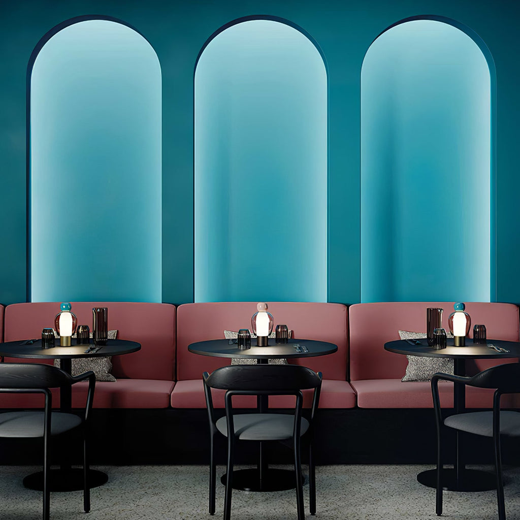 Small Clérachic table lamps glowing on café tables in front of a row of rose-pink booths, under three tall teal arches.