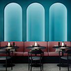 Small Clérachic table lamps glowing on café tables in front of a row of rose-pink booths, under three tall teal arches.