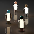 Four lit Clérachic table lamps with pink, brown, turquoise and dark green caps, standing apart on a dark reflective surface.