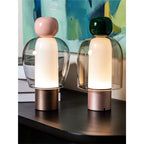 Pink-capped and dark green-capped Clérachic table lamps lit side by side on a black ledge, with a teal painting behind.