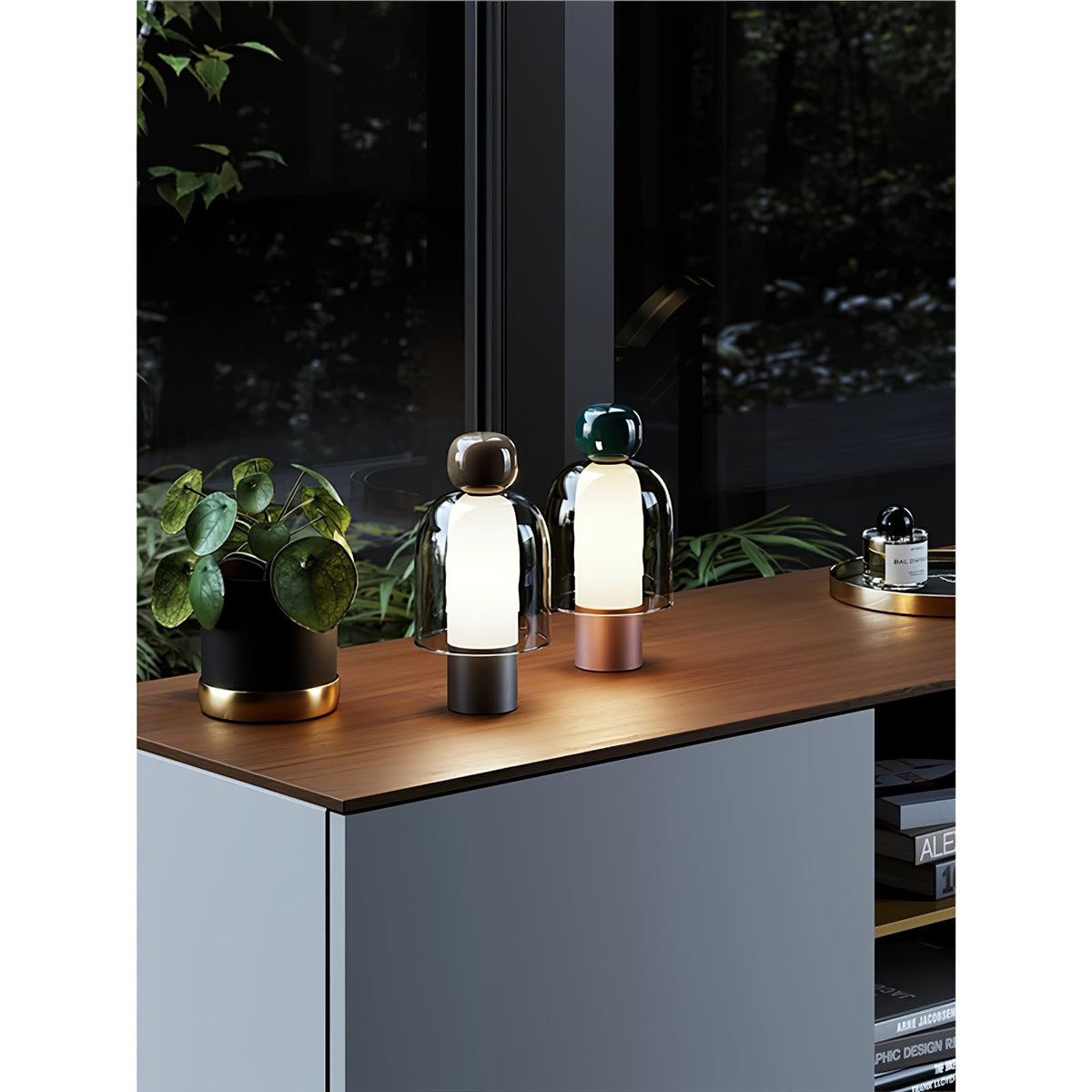 Distant view of two small Clérachic table lamps on a wood-topped cabinet by a window, with a plant in a black pot beside them.