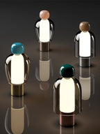 Four tall Clérachic table lamps with pink, blue, brown and dark green caps, glowing on a dark surface, seen from above.