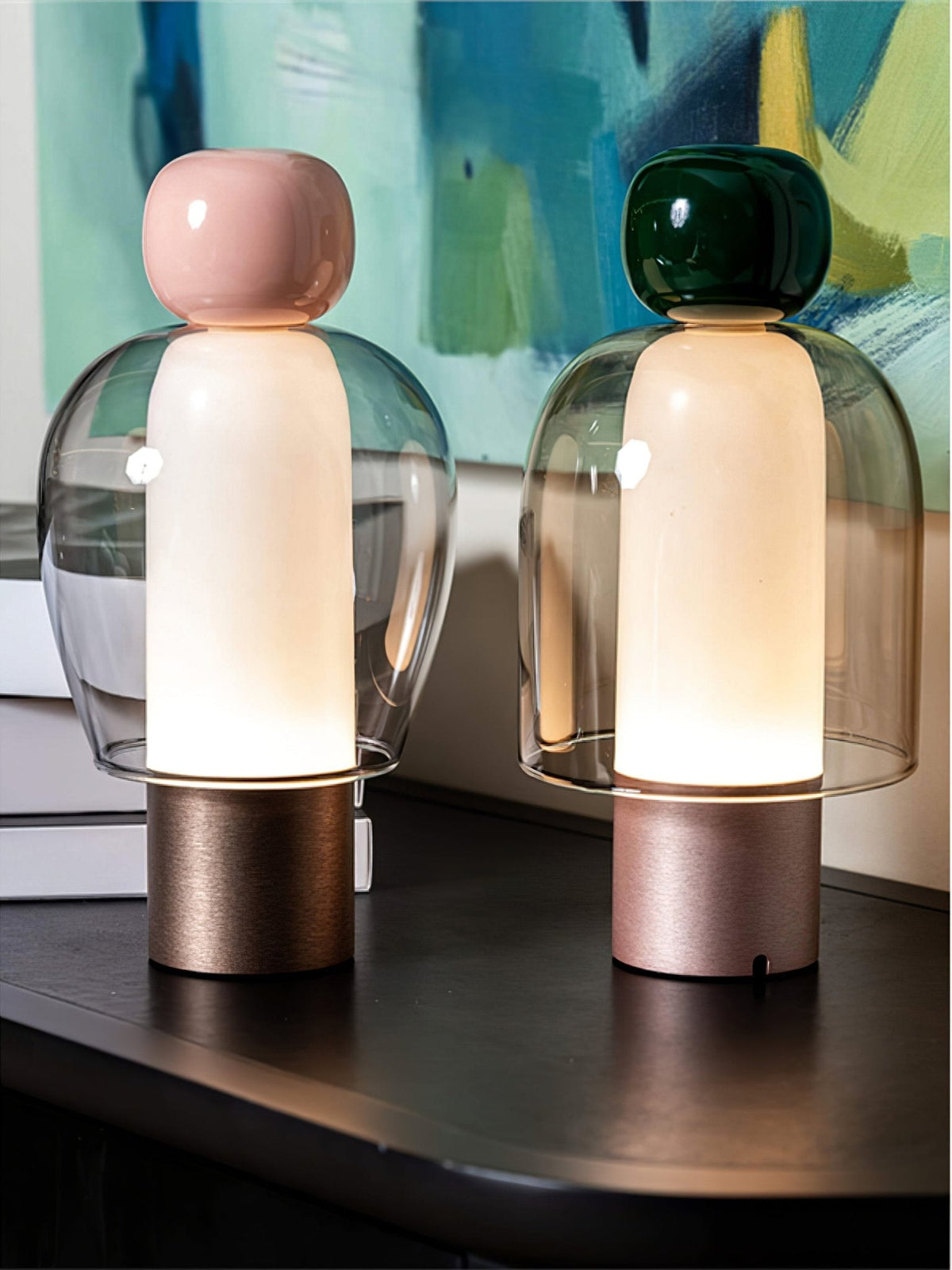 Close view of pink-capped and dark green-capped Clérachic table lamps, their glass domes lit warm, against a teal painting.
