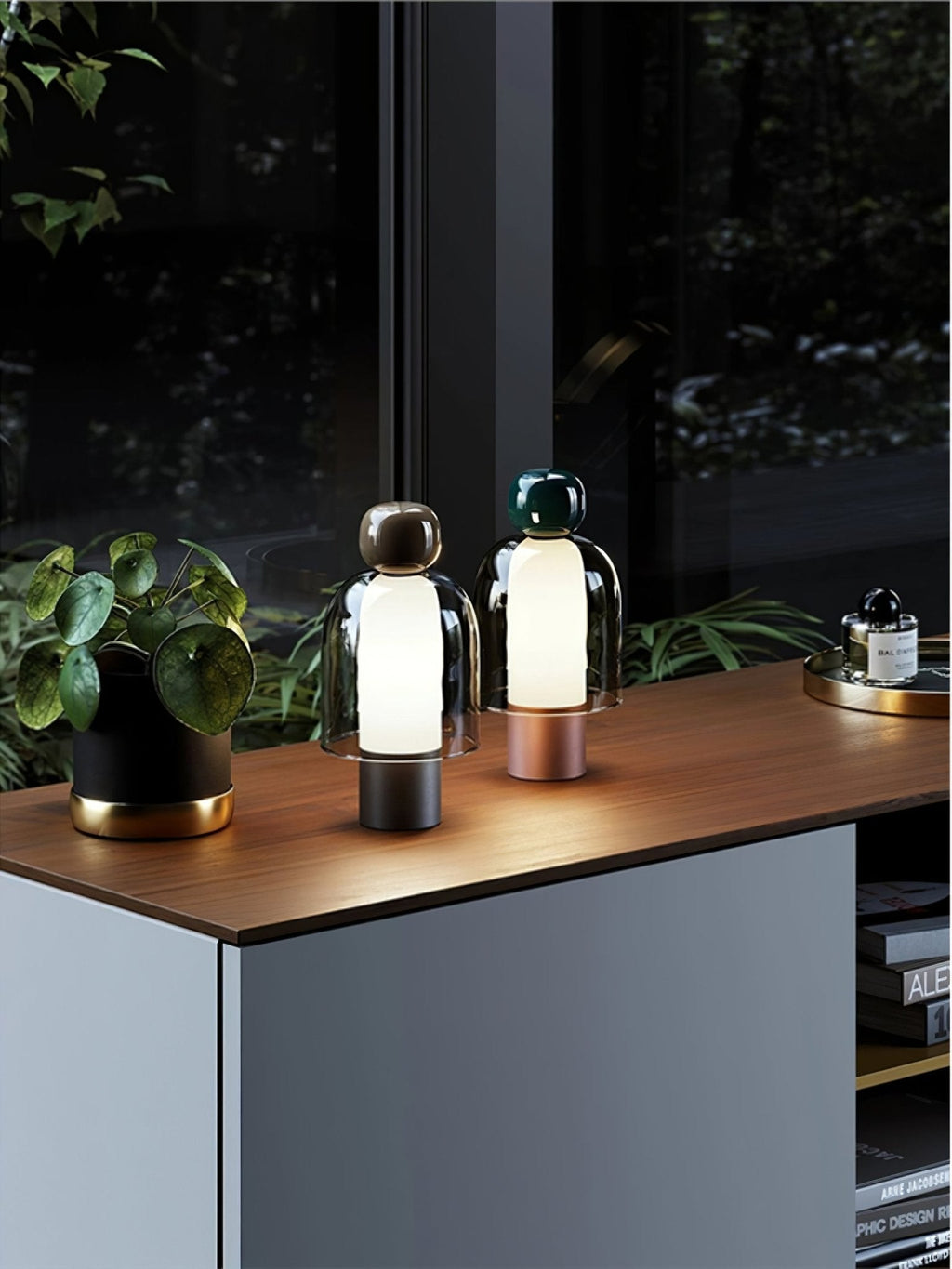 Two small Clérachic table lamps on a wood-topped cabinet near a window, with a plant, a tray and a bottle at the right.