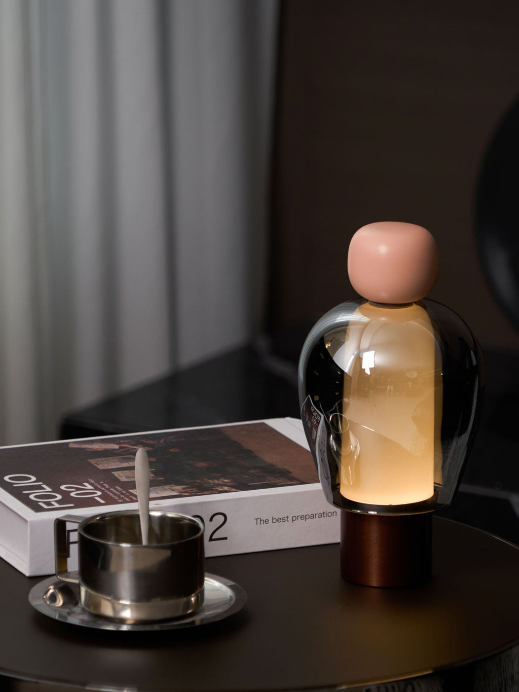 A pink-capped Clérachic table lamp lit warm on a round dark table beside a steel cup and saucer and an open book.