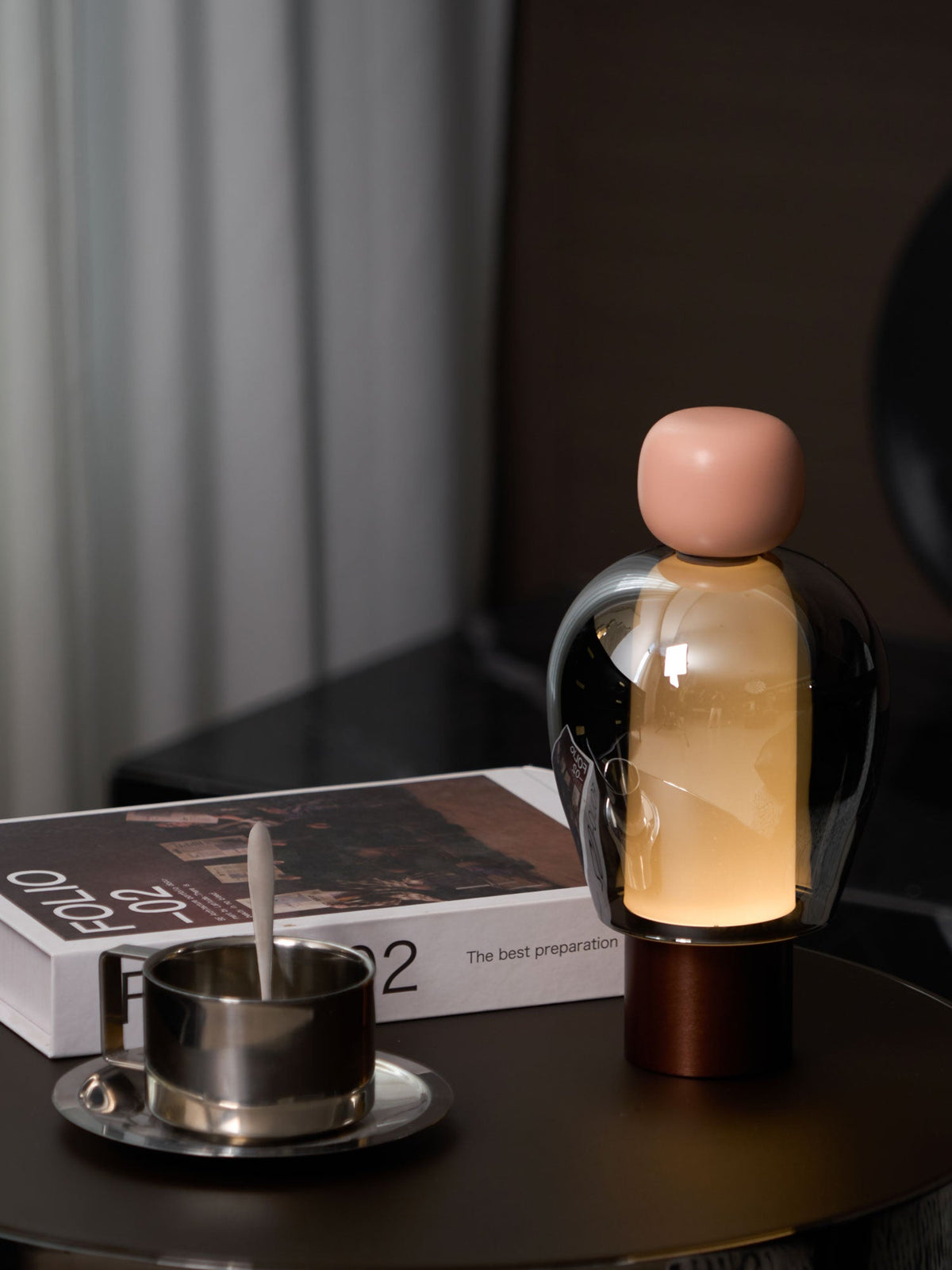 A pink-capped Clérachic table lamp lit warm on a round dark table beside a steel cup and saucer and an open book.