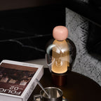 Close view of a pink-capped Clérachic table lamp, its glass dome showing the bulb inside, beside a steel cup and a book.