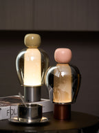 Olive-capped and pink-capped Clérachic table lamps side by side on a round table, with a steel cup and a book in front.