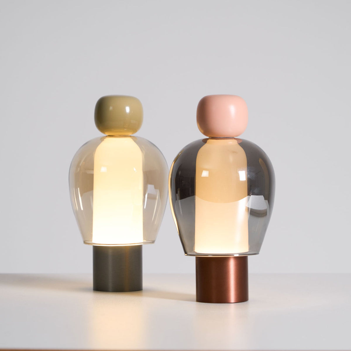 Two lit Clérachic table lamps on a pale surface, the left with an olive cap and steel base, the right pink with a copper base.