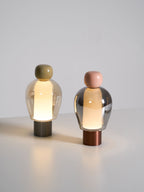 Smaller view of two Clérachic table lamps lit on a pale surface, one olive-capped and one pink-capped, with soft light on the floor.