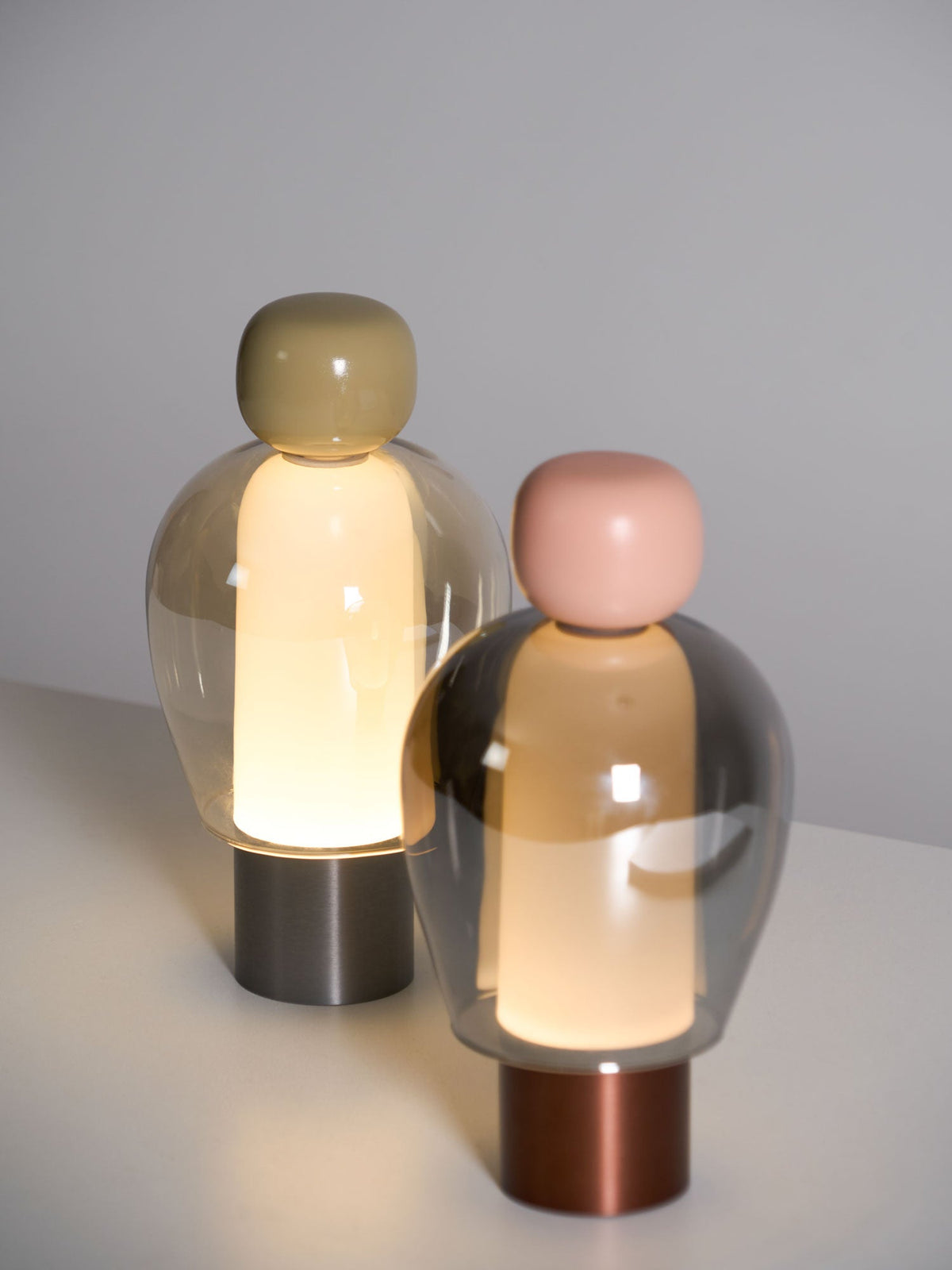 Angled view of two overlapping Clérachic table lamps on a pale surface, the pink-capped one in front of an olive-capped one.