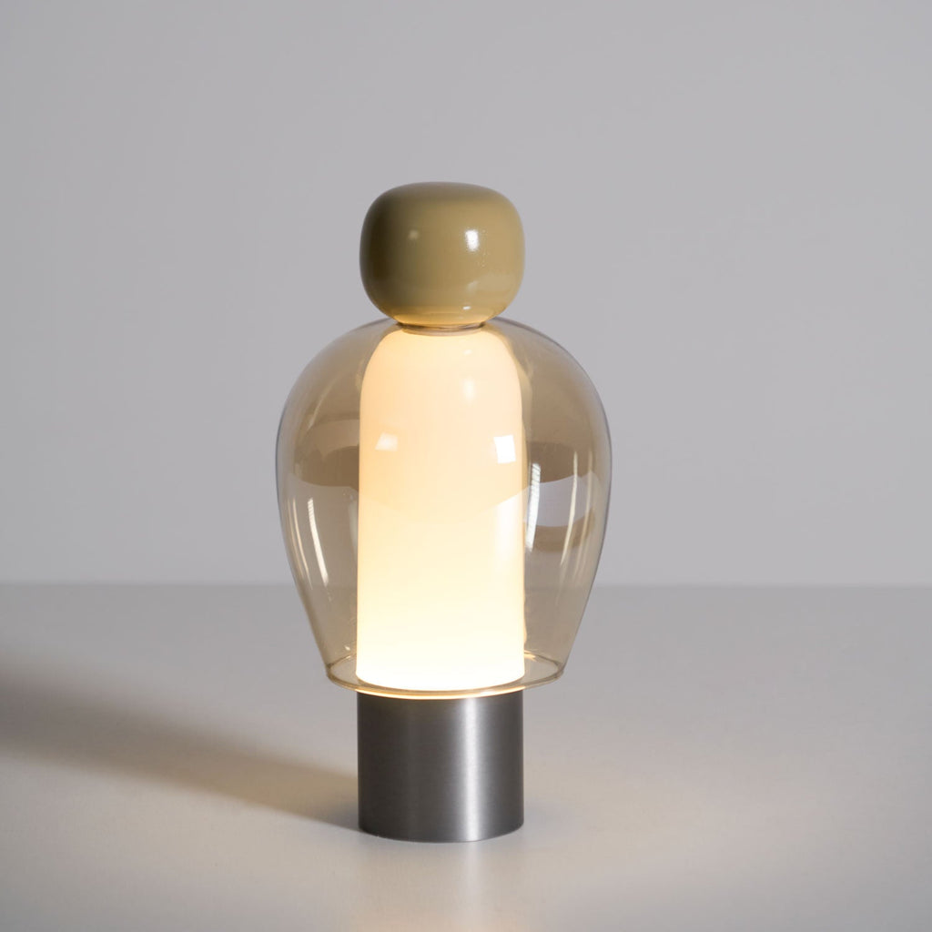 Lit Clérachic table lamp on a pale grey surface, an olive cap above an amber glass dome and a brushed steel base.