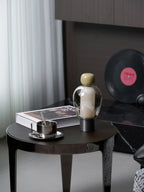 Clérachic table lamp with an olive cap on a round dark table beside a steel cup, a book and a vinyl record, near sheer curtains.