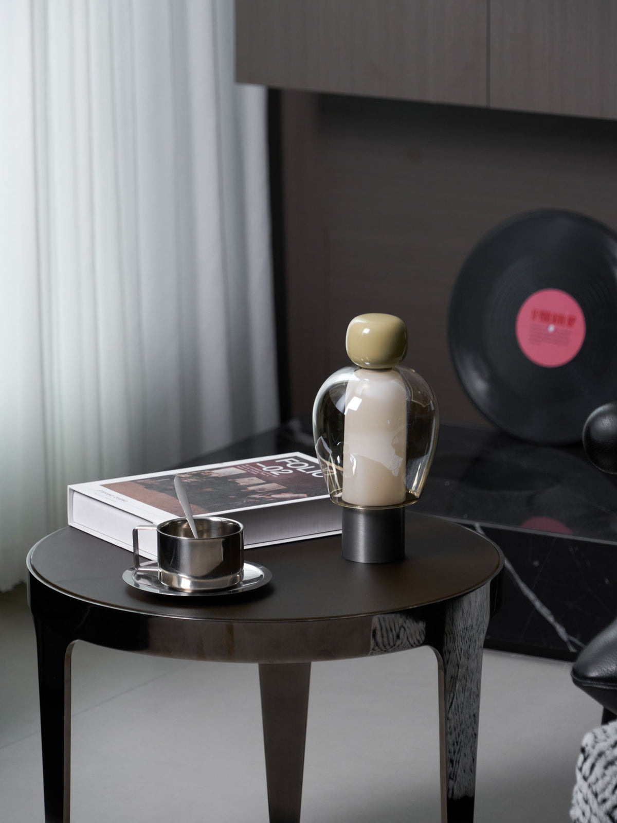 Clérachic table lamp with an olive cap on a round dark table beside a steel cup, a book and a vinyl record, near sheer curtains.