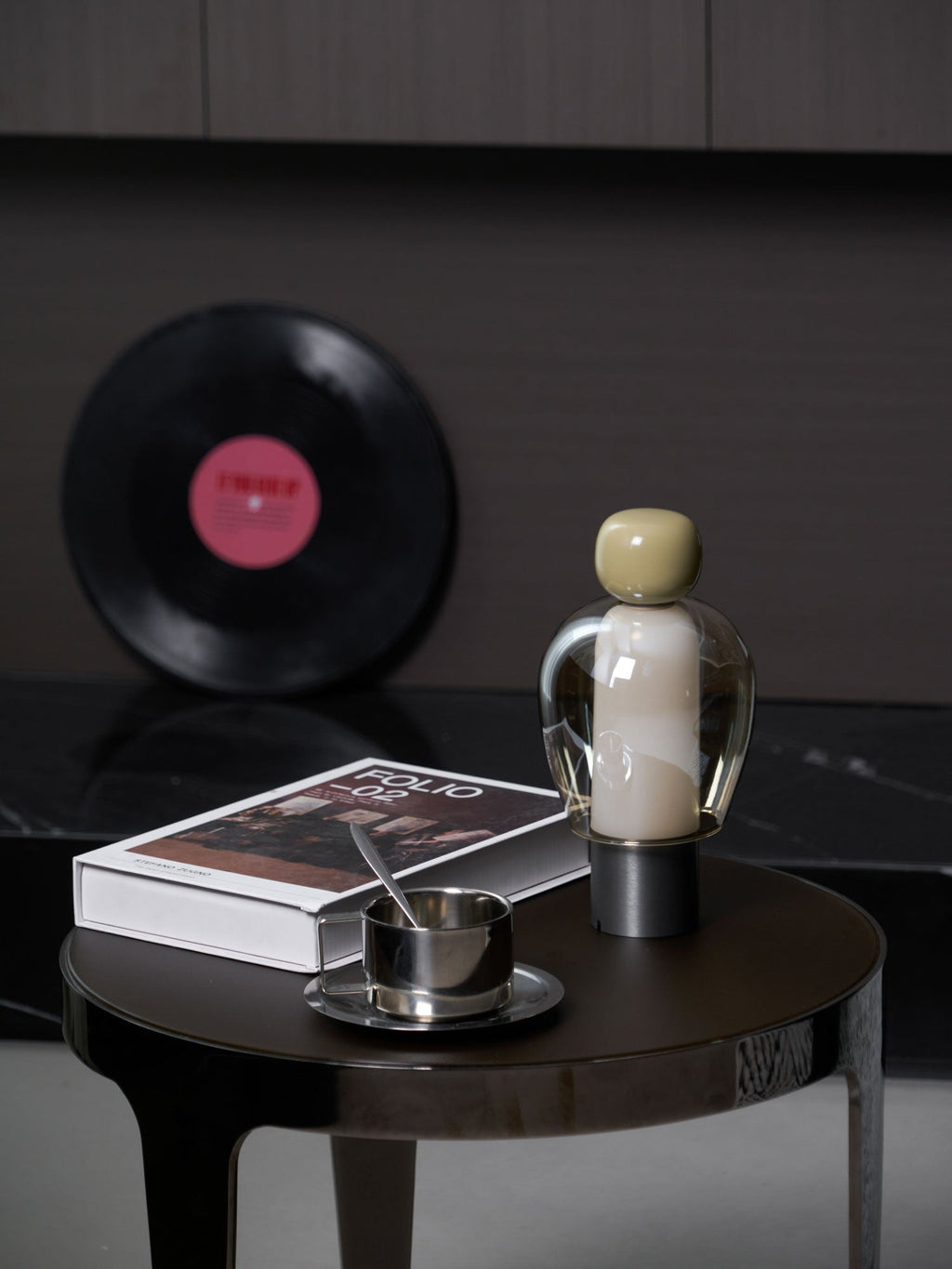 Olive-capped Clérachic table lamp lit softly on a round table, with a steel cup and saucer, a book and a record leaning behind.