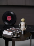 Olive-capped Clérachic table lamp lit softly on a round table, with a steel cup and saucer, a book and a record leaning behind.