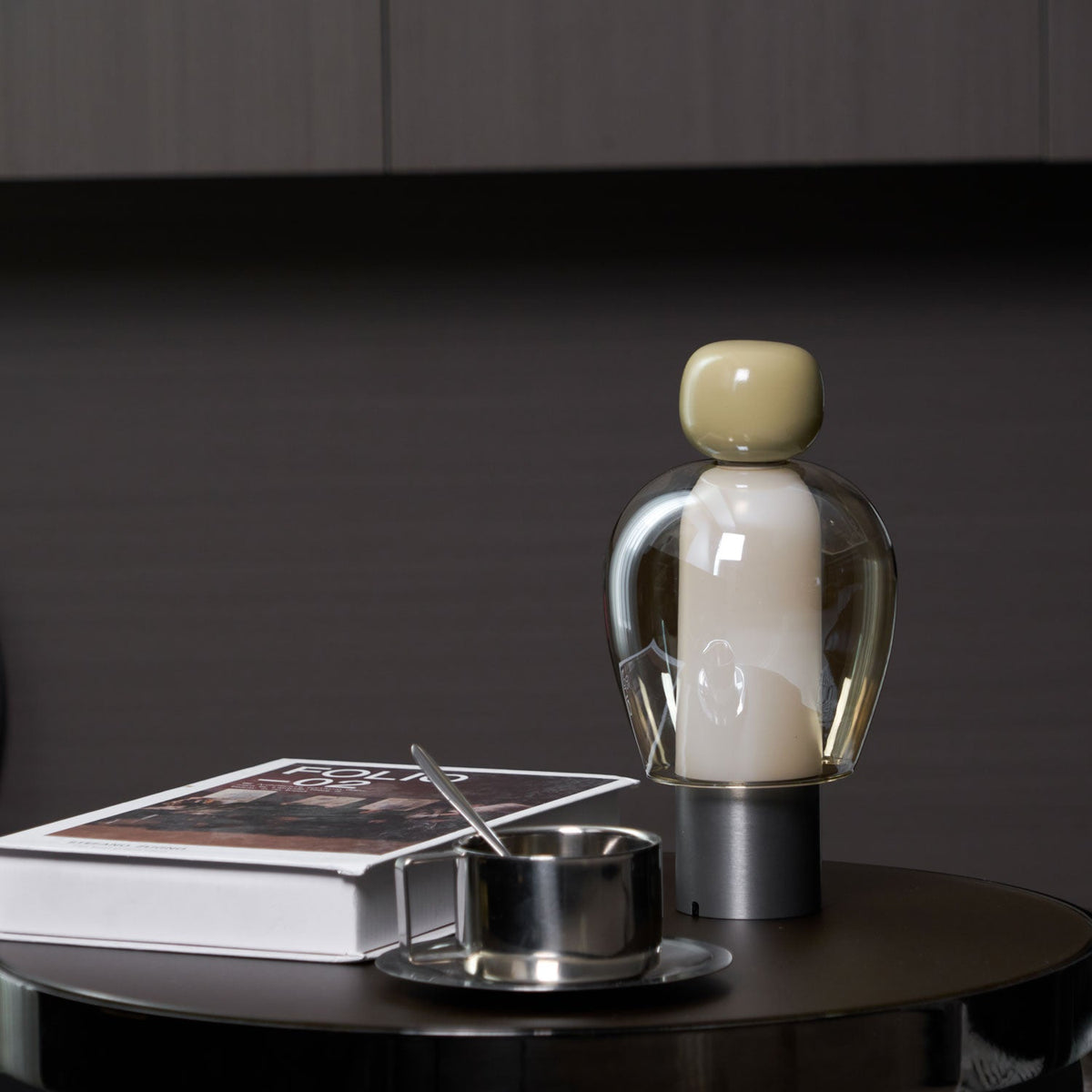 Near view of an olive-capped Clérachic table lamp beside a steel cup and a white book on a dark round table.