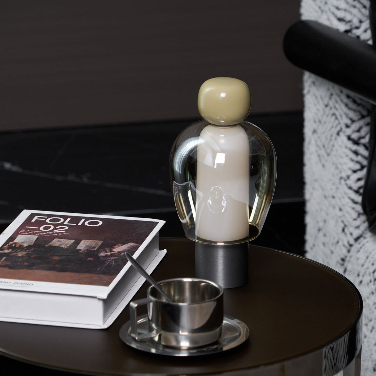 Olive-capped Clérachic table lamp on a dark round table, with a steel cup and spoon and a book, and a black marble wall behind.
