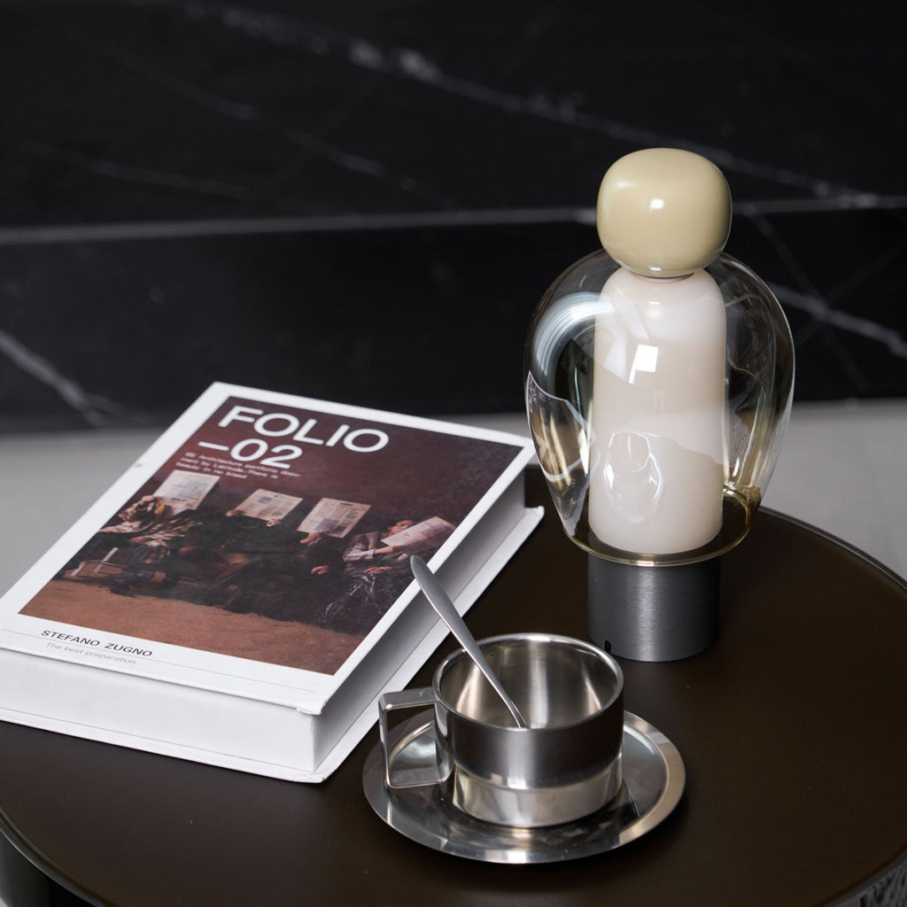 Close, slightly raised view of an olive-capped Clérachic table lamp, a steel cup and saucer and a book on a dark table.