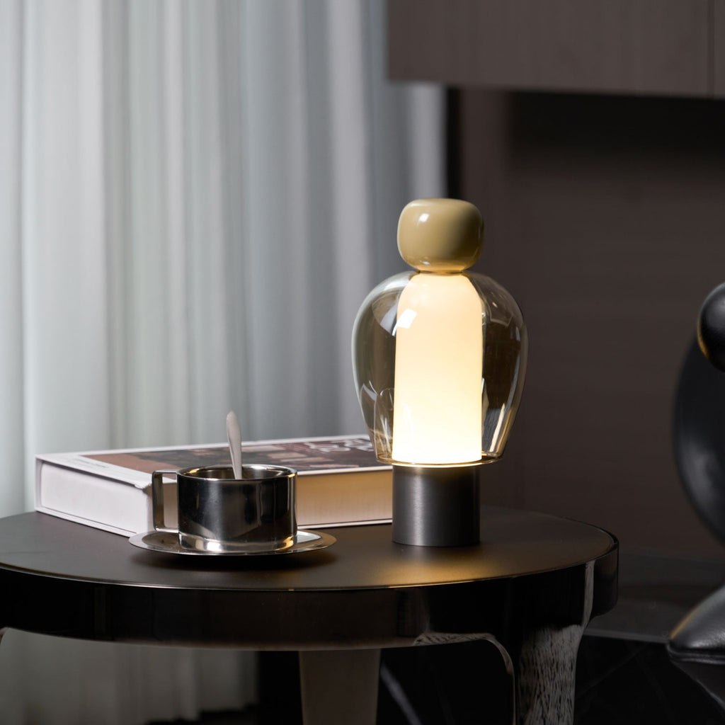 Lit, olive-capped Clérachic table lamp on a dark glossy table, with a steel cup and a book, and sheer curtains at the left.