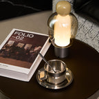 Glowing Clérachic table lamp with an olive cap and a steel base, set beside a book and a steel cup on a dark round table.