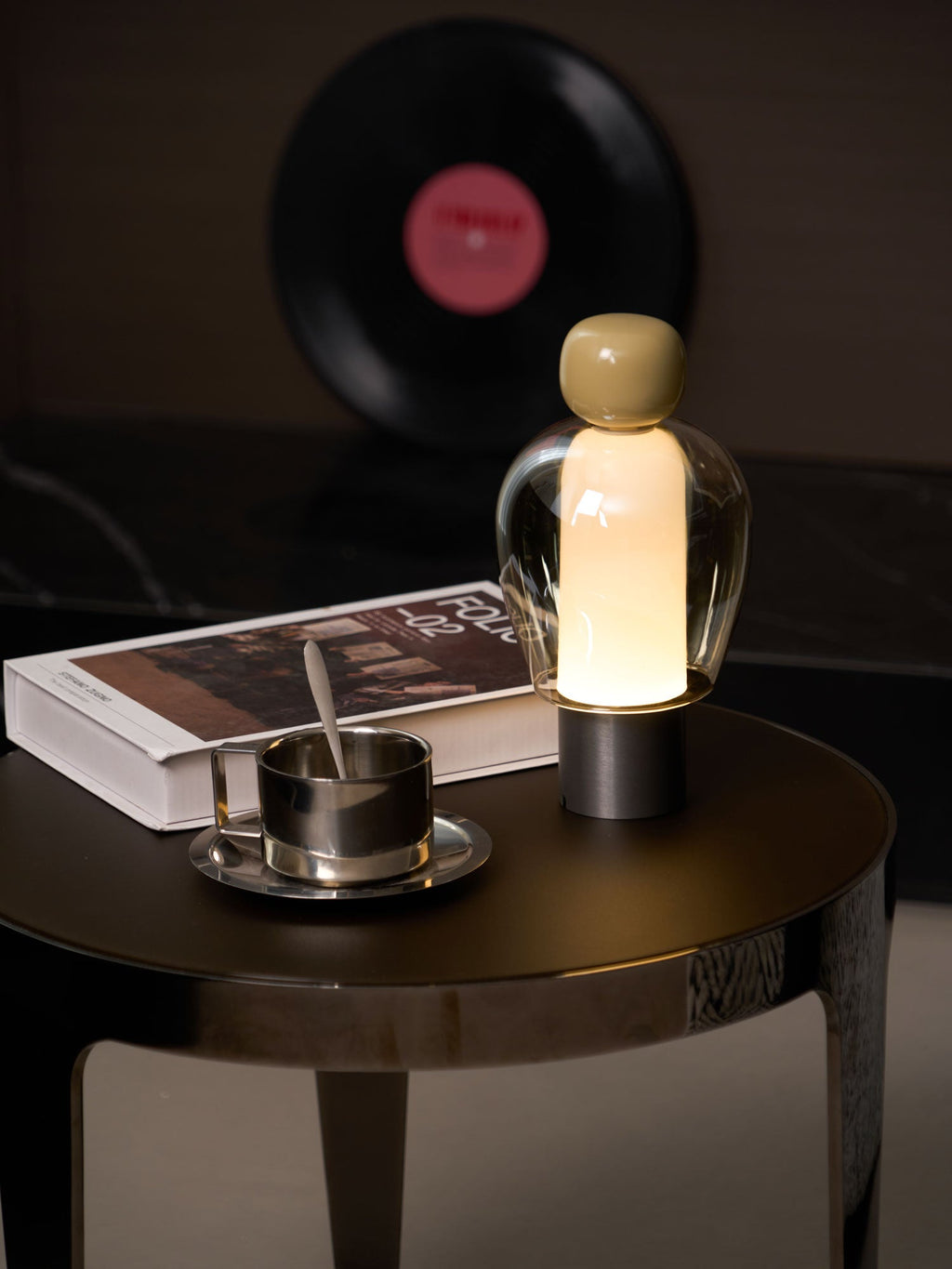 A lit Clérachic table lamp with an olive cap, on a round table with a book and steel cup, and a record on the wall behind.