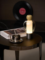 A lit Clérachic table lamp with an olive cap, on a round table with a book and steel cup, and a record on the wall behind.
