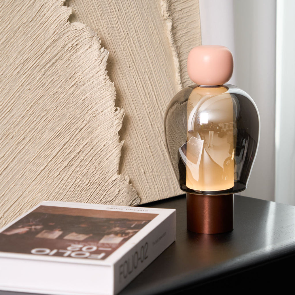 Pink-capped Clérachic table lamp with a smoked glass dome and copper base, beside a large book and a ridged plaster panel.