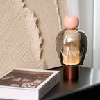 Pink-capped Clérachic table lamp with a smoked glass dome and copper base, beside a large book and a ridged plaster panel.