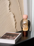 A pink-capped Clérachic table lamp on a black table, seen from further back, with a book at the front and ridged plaster behind.