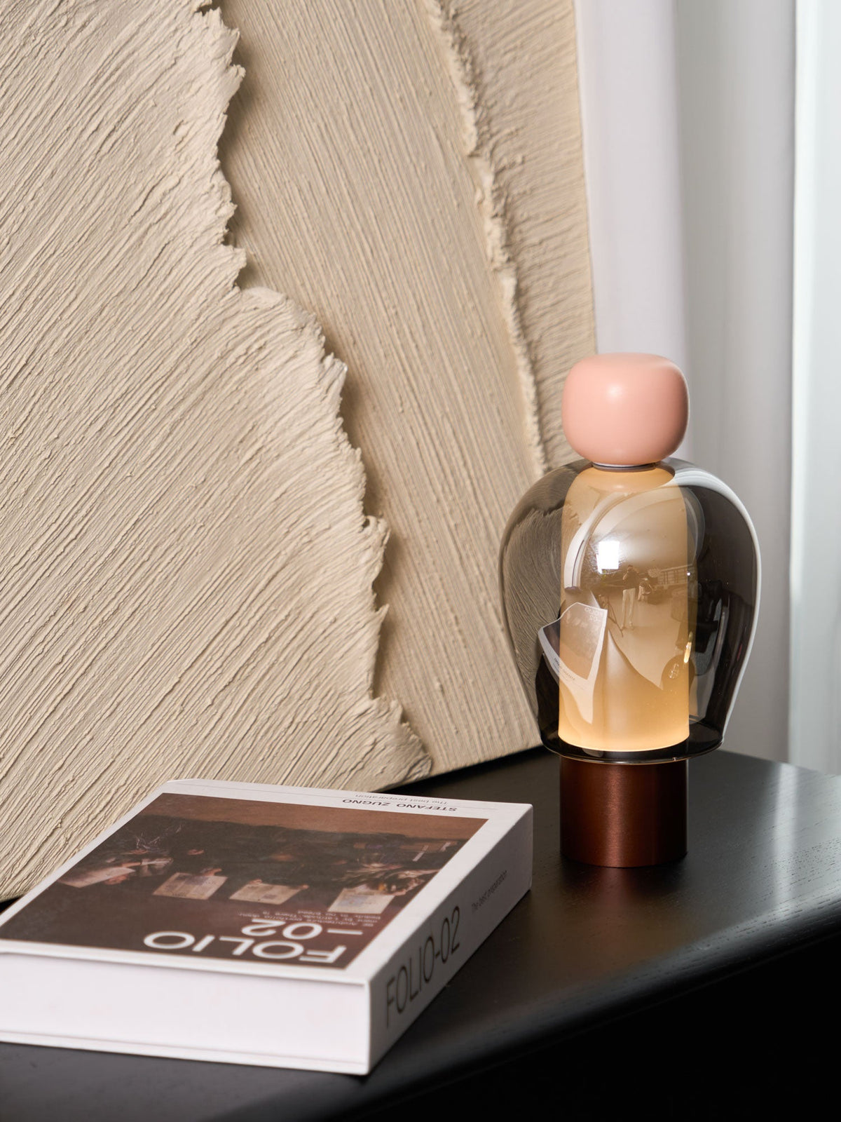 A pink-capped Clérachic table lamp on a black table, seen from further back, with a book at the front and ridged plaster behind.