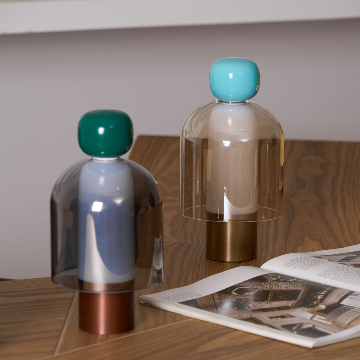Green-capped and turquoise-capped Clérachic table lamps on a herringbone wood desk, beside an open magazine and a pale wall.