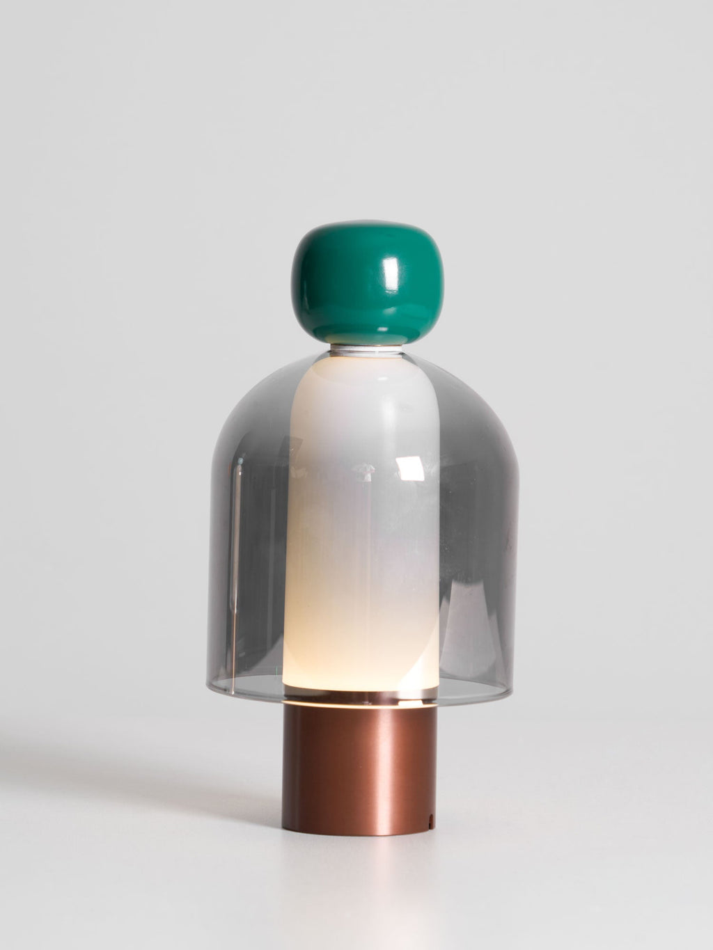 Lit Clérachic table lamp on a pale backdrop, a green cap above a grey glass dome with a soft glow inside and a copper-toned base.