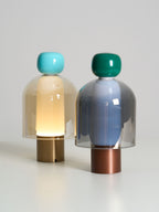 Two lit Clérachic table lamps on a pale surface, one with a turquoise cap and a brass base, one with a green cap and a copper base.