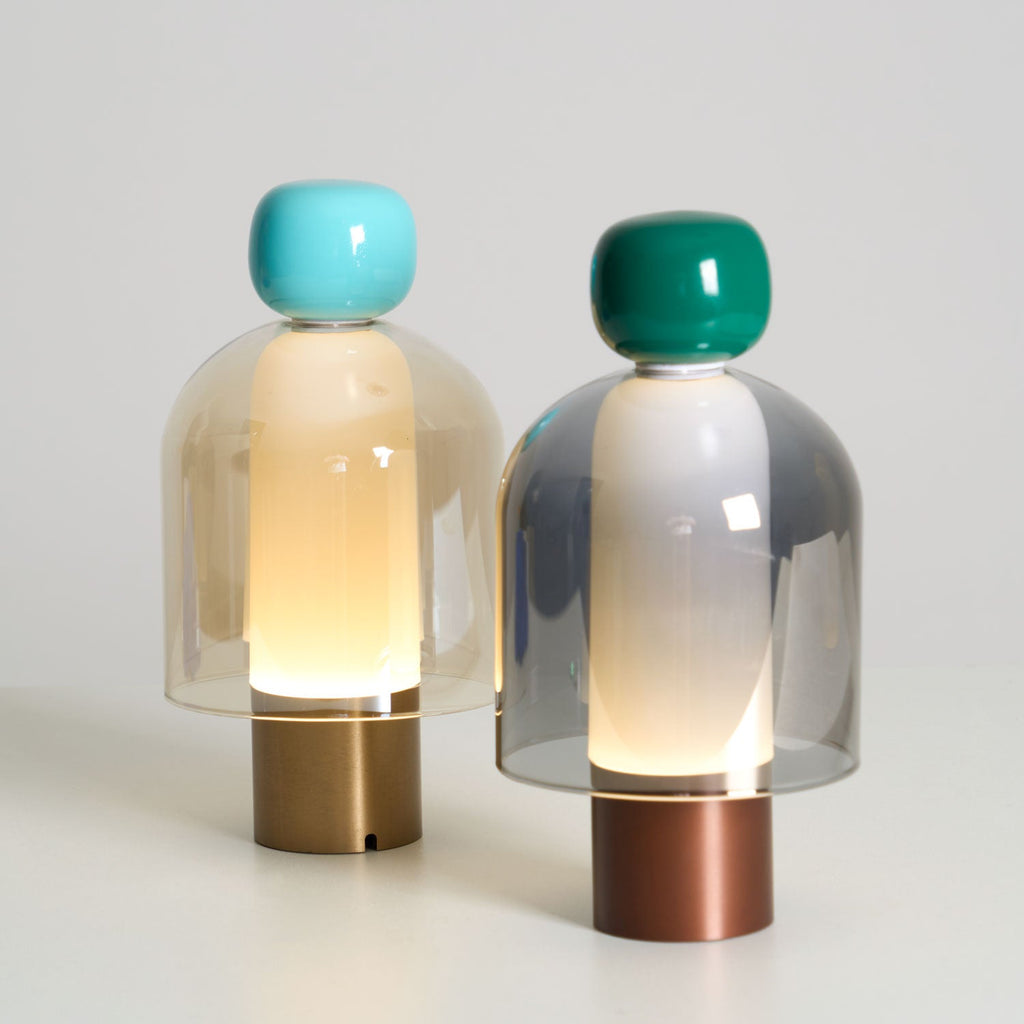 Close view of a turquoise-capped Clérachic table lamp glowing beside a green-capped one with a grey dome, on a pale surface.