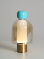 Lit Clérachic table lamp with a turquoise cap and an amber glass dome on a brass base, against a soft grey background.