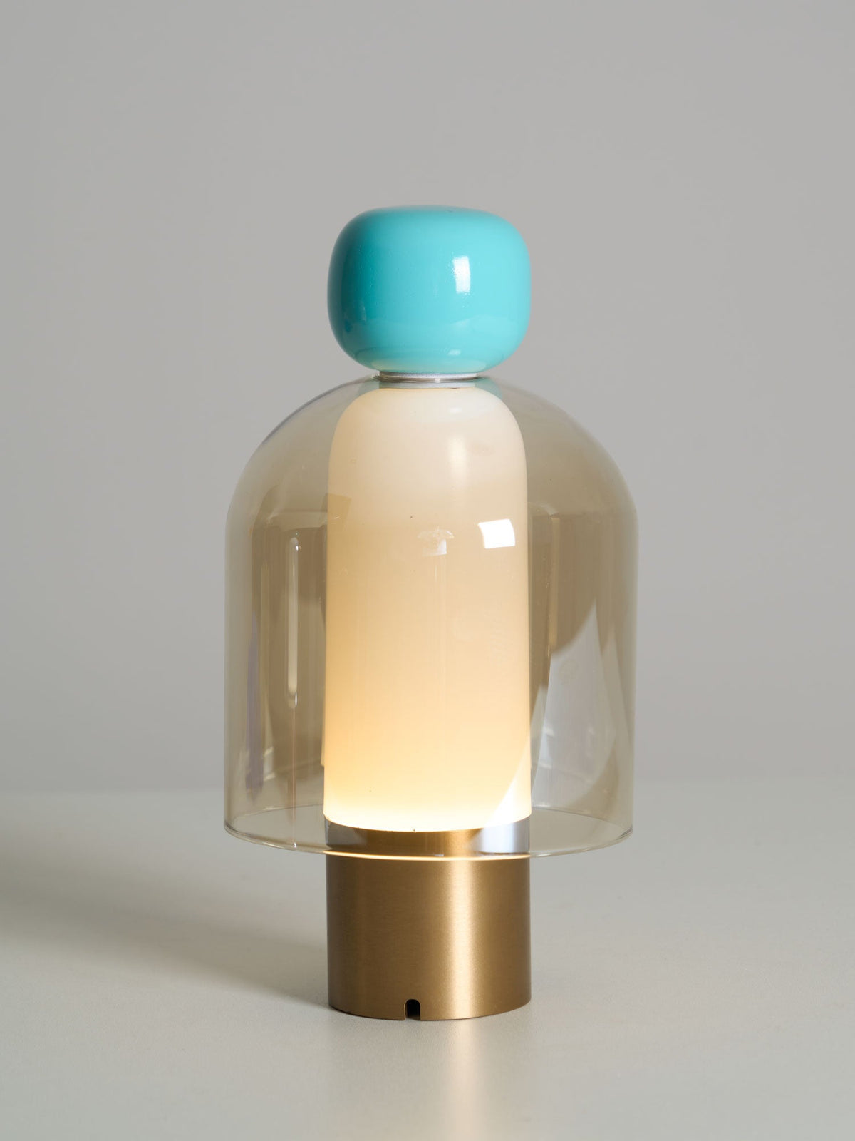 Lit Clérachic table lamp with a turquoise cap and an amber glass dome on a brass base, against a soft grey background.