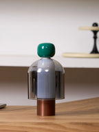 Green-capped Clérachic table lamp with a smoked glass dome and copper base on a wood surface, a pale shelf and a dark stand behind.