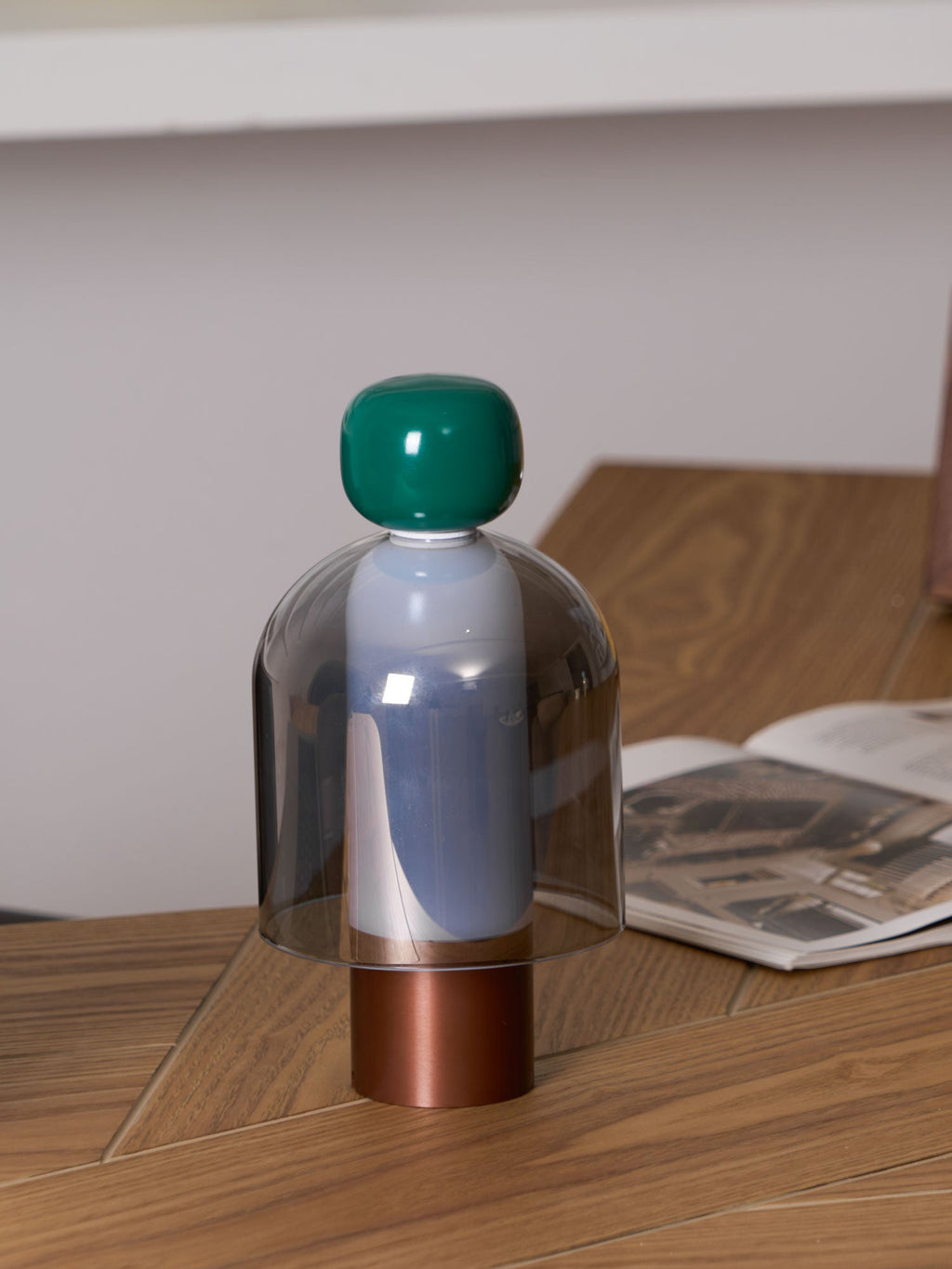 A green-capped Clérachic table lamp on a herringbone wood desk beside an open magazine, with a pale wall behind.