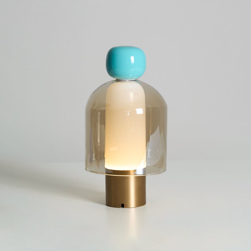 Turquoise-capped Clérachic table lamp on a brass base, its amber glass dome glowing gently against a soft grey wall.