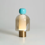 Turquoise-capped Clérachic table lamp on a brass base, its amber glass dome glowing gently against a soft grey wall.