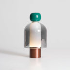 Lit Clérachic table lamp on white, a green cap over a grey glass dome with a glowing inner column and a copper-toned base.